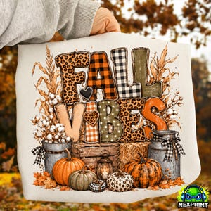 May include: A white sweatshirt featuring a graphic print with the text FALL VIBES. The letters are filled with leopard print, orange and black plaid, and buffalo check patterns. Below are pumpkins, a metal milk can, a lantern, and cotton stems.
