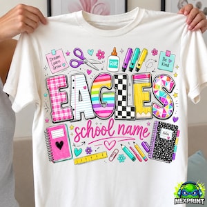 Könnte beinhalten: Ein weißes T-Shirt mit einer bunten Grafik, die das Wort EAGLES in großen, gemusterten Buchstaben zeigt. Das Design enthält Schulmaterialien wie Schere, Kleber, Stifte, ein Lineal und Notizbücher sowie den Text Dream Learn Grow und Be Kind.