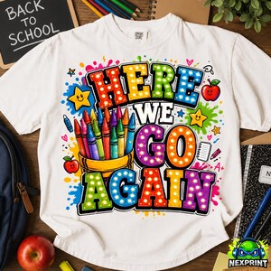 May include: A white t-shirt featuring a colorful graphic with the text "HERE WE GO AGAIN" in a dotted, multi-colored font. The design includes a cup filled with various colored crayons, two red apples, yellow stars, and paint splatters.