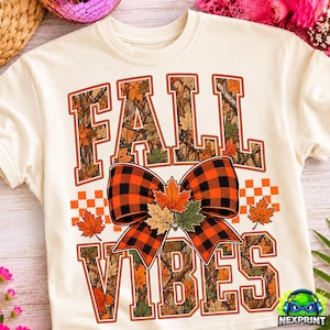 May include: A cream-colored t-shirt featuring the text FALL VIBES in block letters filled with a camouflage and autumn leaf pattern. A large red and black buffalo plaid bow sits between the words, accented with three maple leaves.