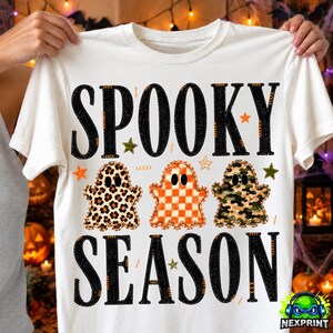 May include: A white t-shirt featuring the black text SPOOKY SEASON. Between the words are three ghost illustrations: one with a leopard print, one with an orange and white checkerboard pattern, and one with a green and brown camouflage pattern.