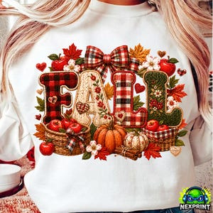 Op de afbeelding: Een wit sweatshirt met een grafische print van het woord FALL in grote, textuurrijke letters. De letters zijn versierd met rood-zwarte ruiten, crèmekleurig breiwerk en groene bloemmotieven. Pompoenen, appels en bladeren omringen de tekst.