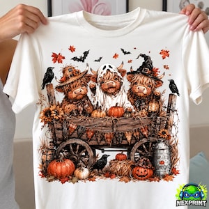 Può includere: Una t-shirt bianca con una grafica di tre mucche Highland su un carro di legno. Una mucca indossa un cappello, una è vestita da fantasma e una indossa un cappello da strega. La scena include zucche, corvi neri, pipistrelli e foglie autunnali.