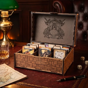 May include: A wooden card storage box with a stone-patterned exterior and a hinged lid featuring an engraved shield emblem with the word QUEST. The interior contains multiple dividers holding various illustrated game cards.