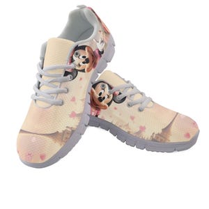 K&ouml;nnte beinhalten: Paar beige Sportschuhe mit einem Cartoon-Charakter-Design. Die Schuhe haben eine hellgraue Sohle, hellgraue Schn&uuml;rsenkel und ein sich wiederholendes Muster aus rosa Herzen. Die Cartoonfigur tr&auml;gt eine rosa Schleife.
