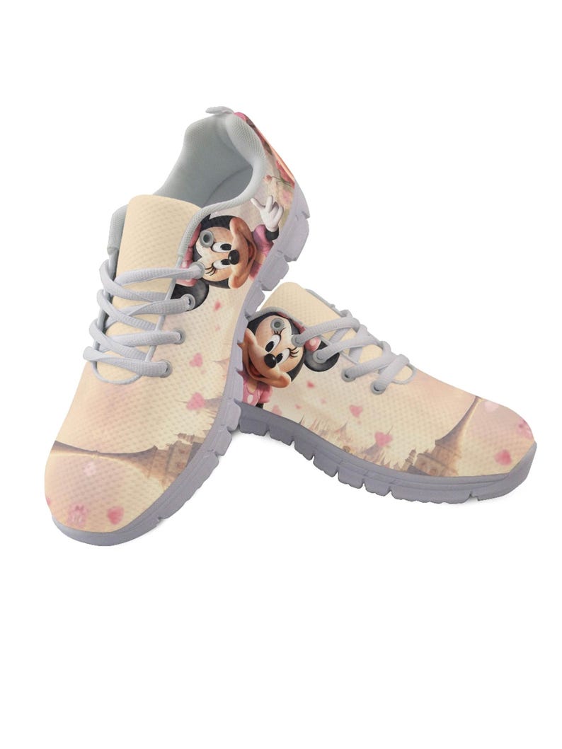 K&ouml;nnte beinhalten: Paar beige Sportschuhe mit einem Cartoon-Charakter-Design. Die Schuhe haben eine hellgraue Sohle, hellgraue Schn&uuml;rsenkel und ein sich wiederholendes Muster aus rosa Herzen. Die Cartoonfigur tr&auml;gt eine rosa Schleife.