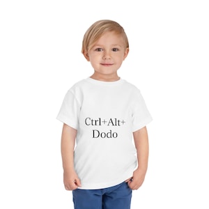 Peut inclure: Un t-shirt blanc pour enfant avec le texte "Ctrl+Alt+Dodo" en noir. Le t-shirt à manches courtes est porté par un jeune enfant souriant à la caméra. L'enfant porte un pantalon bleu.