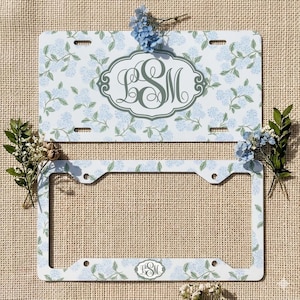May include: A white license plate and license plate frame with a blue floral pattern. The license plate features a monogram in a decorative oval. The frame and plate have a matching design with a light blue hydrangea pattern.