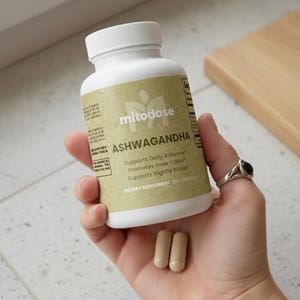 Puede incluir: Una botella blanca de suplemento diet&eacute;tico "mitodose ASHWAGANDHA", sostenida en una mano. La etiqueta es de color beige con texto verde, y la botella tiene una tapa blanca. Dos c&aacute;psulas est&aacute;n en la mano. El texto en la etiqueta dice "Supports Daily Balance, Promotes Inner Calm, Supports Nightly Ritual".