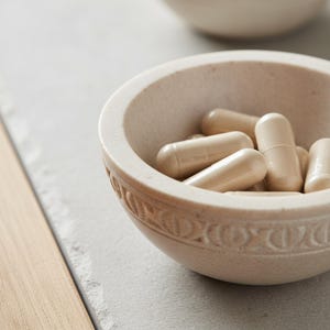 Puede incluir: Un cuenco de piedra beige tallada lleno de varias c&aacute;psulas beige. El cuenco tiene un borde decorativo y se encuentra sobre una superficie gris claro. Otro cuenco similar es ligeramente visible en el fondo.