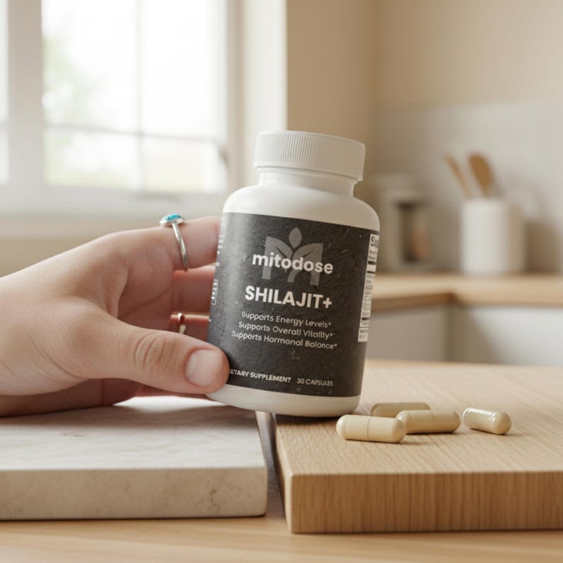 May include: A white bottle of "SHILAJIT+" dietary supplement with a black label, held by a hand. The label includes the brand name "mitodose" and the text "Supports Energy Levels, Overall Vitality, and Hormonal Balance." Several capsules are on a wooden surface.