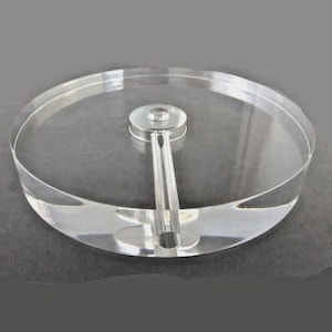 May include: A clear acrylic cake stand with a silver metal handle. The stand has a circular base and a raised edge. The handle is attached to the center of the base and extends upwards.