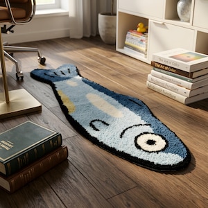 Pode incluir: Um tapete em forma de peixe em tons de azul, branco e amarelo, com uma borda preta, colocado sobre um chão de madeira. O design do tapete adiciona um toque lúdico ao ambiente. As dimensões do tapete não são visíveis.