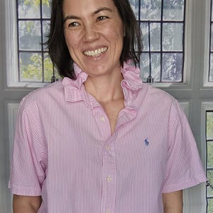 Peut inclure: Une chemise à manches courtes boutonnée avec un motif à rayures verticales roses et blanches, un col à volants et un petit logo bleu brodé sur la poitrine gauche. La chemise présente un ourlet arrondi et est portée avec une jupe beige unie.