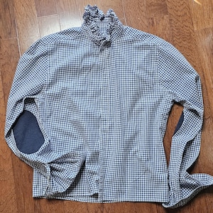 May include: A long-sleeved button-down shirt featuring a blue and white gingham check pattern. The shirt includes a high ruffled collar and solid dark blue elbow patches on each sleeve, providing a distinct contrast to the overall checkered design.