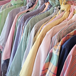 Può includere: Una collezione di camicie a maniche lunghe con bottoni appese a un appendiabiti in metallo. Le camicie presentano vari motivi, tra cui righe, quadri e colori a tinta unita nelle tonalità del giallo, rosa, blu, verde e bianco in cotone.
