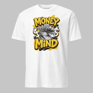 May include: White t-shirt with a graphic design. The design features the words "MONEY ON MIND" in yellow, outlined in black, with dollar signs and a bag of money. The shirt is made of a soft material.