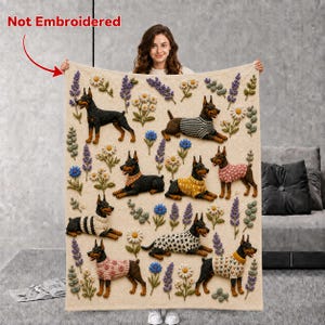 May include: A cream-colored throw blanket featuring a repeating pattern of Doberman dogs wearing various sweaters, interspersed with colorful floral sprigs and lavender stalks. The text "Not Embroidered" appears in red at the top left corner.
