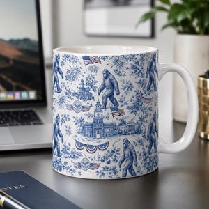 May include: White ceramic mug with a blue toile pattern featuring Bigfoot, American flags, fireworks, and historical buildings. The mug has a white handle and is set on a dark surface.
