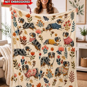 May include: A cream-colored throw blanket featuring a repeating pattern of Australian Shepherd dogs in various poses, some wearing sweaters, interspersed with colorful floral illustrations. A red text box reads NOT EMBROIDERED.