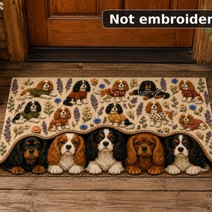 May include: A rectangular doormat featuring a repeating pattern of Cavalier King Charles Spaniels in various sweaters, interspersed with floral and lavender sprigs. The bottom edge shows five spaniels peeking out. Text reads: Not embroidered.