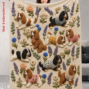 Może przedstawiać: Beżowy koc z powtarzającym się wzorem psów rasy Cavalier King Charles w różnych swetrach, przeplatany ilustracjami lawendy, stokrotek i pomarańczowych kwiatów. Widoczny jest napis Not Embroidered po lewej stronie koca.