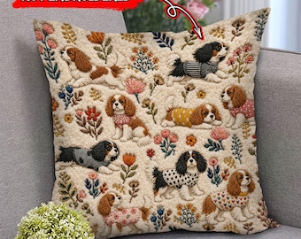 Puede incluir: Un cojín cuadrado con fondo texturizado de color crema que presenta un patrón repetido de perros Cavalier King Charles Spaniel con varios suéteres, intercalados con motivos florales coloridos. El texto NOT EMBROIDERED es visible.