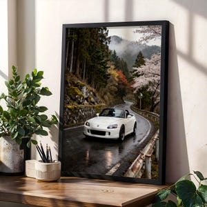 May include: A framed wall art print featuring a white convertible sports car driving on a wet, winding mountain road. The scene includes a dense forest, rocky terrain, and blooming cherry blossom trees under a soft, overcast sky.