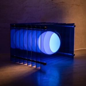 Könnte beinhalten: Eine moderne blaue Lampe mit einer kugelförmigen Lichtquelle. Die Lampe hat eine Reihe vertikaler blauer Paneele, die einen geschichteten Effekt um das zentrale Licht erzeugen. Das Licht strahlt ein weiches, kühles Leuchten aus.