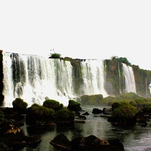 Può includere: Una vasta cascata si getta da una scogliera rocciosa in un fiume scuro. La vegetazione verde e le rocce coperte di muschio circondano la base. Il cielo è nuvoloso, creando un'atmosfera serena.