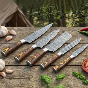 May include: A set of five kitchen knives with patterned steel blades and wooden handles with orange and yellow accents. The knives are arranged on a wooden surface with garlic, tomatoes, and herbs.