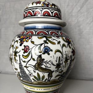 May include: A white ceramic jar with a lid, decorated with vibrant floral and geometric patterns. The jar features a deer illustration and is accented with red, blue, yellow, and green details. The jar is approximately 25 cm tall.