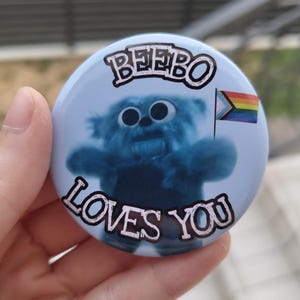 Puede incluir: Una chapa redonda con fondo azul claro que muestra una criatura azul y peluda en el centro. El texto BEEBO LOVES YOU está impreso en negro con un borde blanco. La criatura sostiene una pequeña bandera del orgullo en el lado derecho.