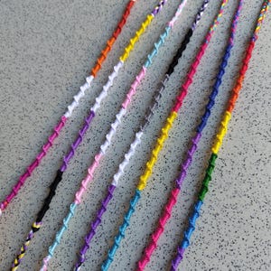 Op de afbeelding: Een collectie van zes dunne, handgemaakte vriendschapsarmbandjes gemaakt van kleurrijk borduurgaren. Elk armbandje heeft een spiraalvormig knooppatroon in diverse levendige tinten, waaronder rood, oranje, geel, groen, blauw, paars, roze, zwart en wit.