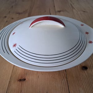 May include: An off-white ceramic serving dish with a matching lid featuring a red arched handle. The lid and base are decorated with thin black concentric lines and small red star-shaped accents arranged in a pattern along the curved edges.