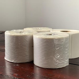 May include: Four rolls of white label paper, wrapped in clear plastic, on a wooden surface.