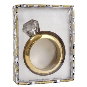 May include: A gold-coloured ring-shaped flask with a clear, faceted gem on top, presented in a decorative box. The flask has a dark brown outer ring and a gold inner ring. The box has a geometric pattern in gold, white, and grey.