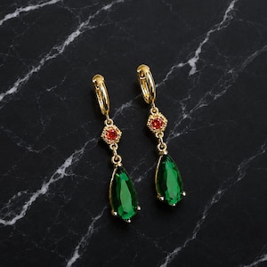 Può includere: Un paio di orecchini pendenti color oro con una piccola gemma rotonda rosso-arancio incastonata in una cornice esagonale testurizzata, collegata a una gemma verde smeraldo più grande, sfaccettata e a forma di goccia.