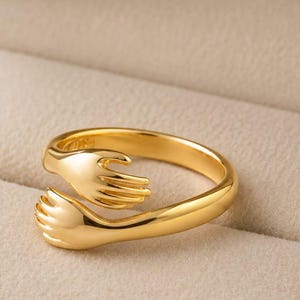 Peut inclure: Une bague dorée dans un écrin à bijoux. La bague présente un motif de deux mains qui s'étreignent. La bague est en métal brillant et réfléchissant. L'écrin est de couleur beige clair.