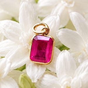 May include: A rectangular pendant featuring a vibrant, deep pink gemstone set in a polished gold-toned metal frame with a small circular bail at the top, resting centrally among a cluster of white jasmine flowers on a scalloped white plate.