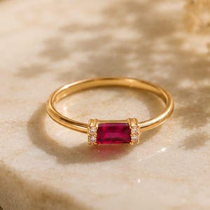 May include: A gold ring featuring a rectangular, deep red gemstone. Small, clear stones are set either side of the red stone. The ring is displayed on a marble surface, with a soft, cream-coloured fabric in the background.