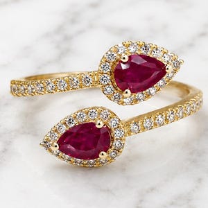 Könnte beinhalten: Ein goldener Bypass-Ring mit zwei birnenförmigen roten Rubinen, die jeweils von einem Kranz aus kleinen weißen Diamanten umgeben sind. Die Ringschiene ist ebenfalls mit kleinen weißen Diamanten besetzt, was ein funkelndes, offenes Design ergibt.
