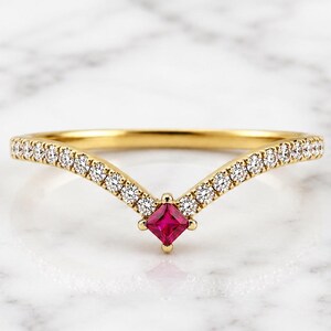 Peut inclure: Une bague en or en forme de V avec un rubis rouge taille princesse au centre, entouré d'une rangée de petits diamants ronds sertis pavé le long de l'anneau, offrant un design symétrique et délicat avec une finition métallique polie.