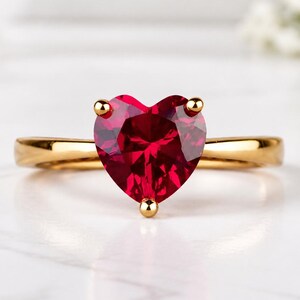 Può includere: Un anello in oro con una prominente pietra preziosa rossa a forma di cuore montata su una montatura a tre griffe. La fascia è liscia e lucida, mettendo in risalto la vibrante pietra cremisi che funge da punto focale del gioiello.