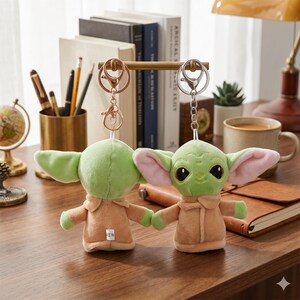 May include: Two plush green character keychains with large ears and tan robes. The keychains are attached to metal rings. The background includes books, a coffee mug, and a desk lamp.