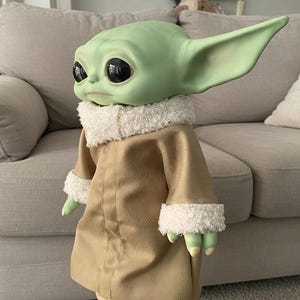 May include: A green-skinned doll with large black eyes and pointed ears, wearing a tan coat with white fluffy trim. The doll's hands and feet are green, and it is posed in front of a light-coloured sofa.