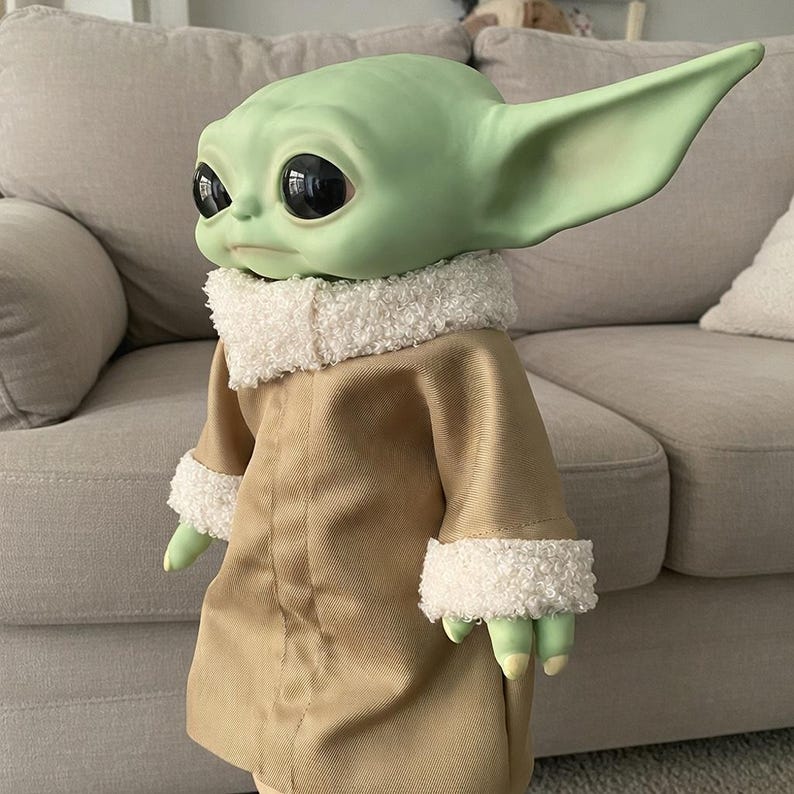 May include: A green-skinned doll with large black eyes and pointed ears, wearing a tan coat with white fluffy trim. The doll's hands and feet are green, and it is posed in front of a light-coloured sofa.