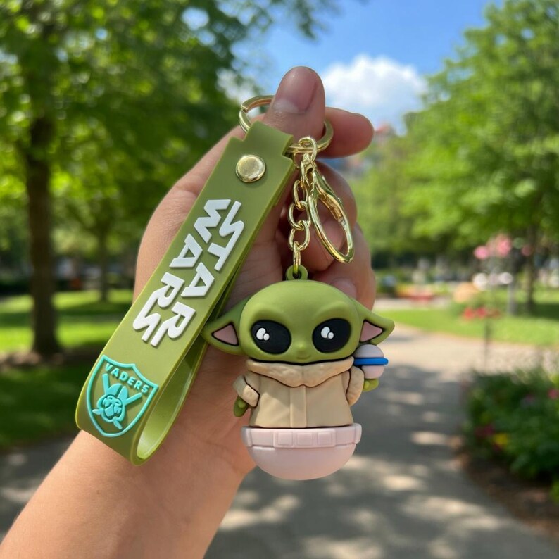 Puede incluir: Un llavero de silicona verde con una figura del personaje Grogu y una correa a juego con el texto blanco STAR WARS y un logotipo VADERS azul claro. El llavero incluye una anilla de metal dorado y un cierre de mosquet&oacute;n.