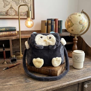 May include: A dark grey penguin-shaped crossbody bag with a white face, yellow feet, and blue accents. The bag has a long strap and is on a stack of books. A desk lamp, globe, and coffee cup are in the background.
