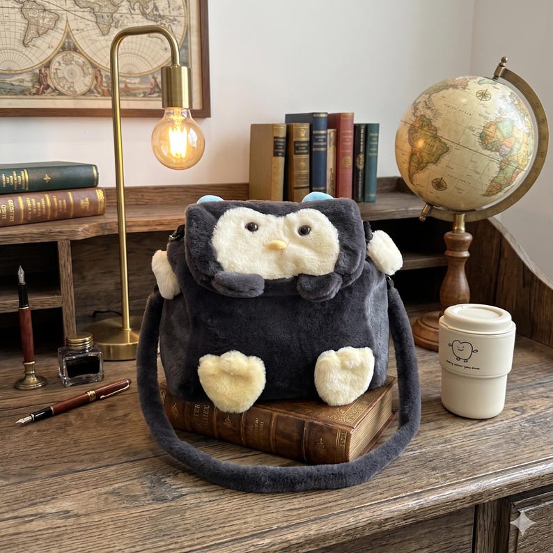 May include: A dark grey penguin-shaped crossbody bag with a white face, yellow feet, and blue accents. The bag has a long strap and is on a stack of books. A desk lamp, globe, and coffee cup are in the background.